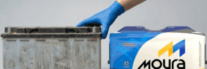 Mão com luva azul segurando uma bateria de carro usada ao lado de uma bateria Moura nova na embalagem.