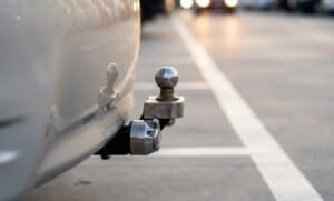 Traseira de um veículo com o engate de reboque e a tomada elétrica visíveis, com o fundo desfocado de um estacionamento.