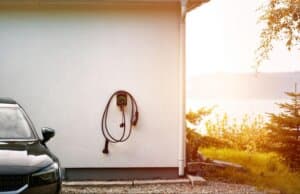 Carregador de carro elétrico instalado na parede externa de uma casa branca, ao lado de um veículo preto.
